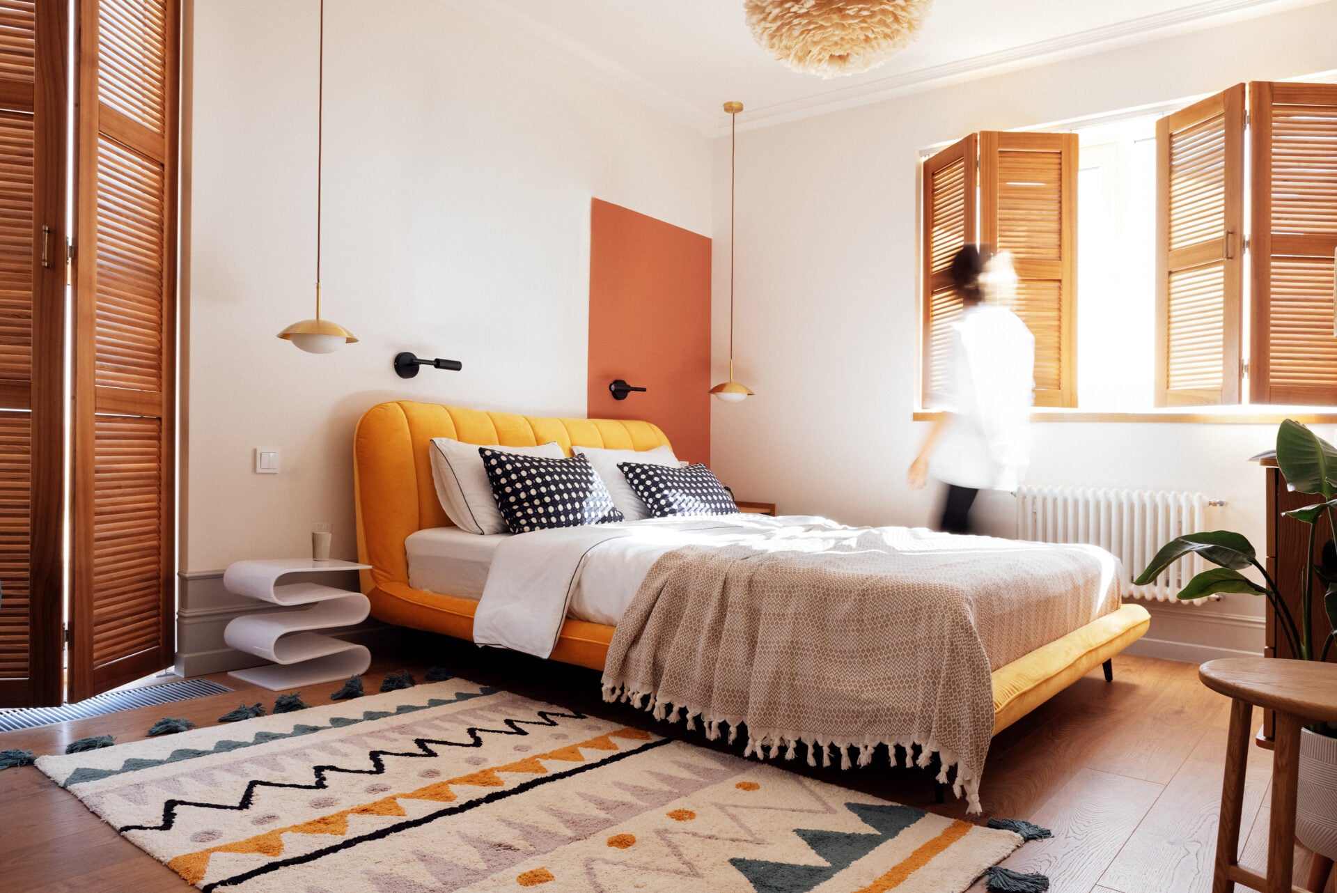 Contemporary bedroom showcases a vibrant yellow bed adorned with decorative pillows, illuminated by sunlight through wooden shutters, enhancing the inviting ambiance