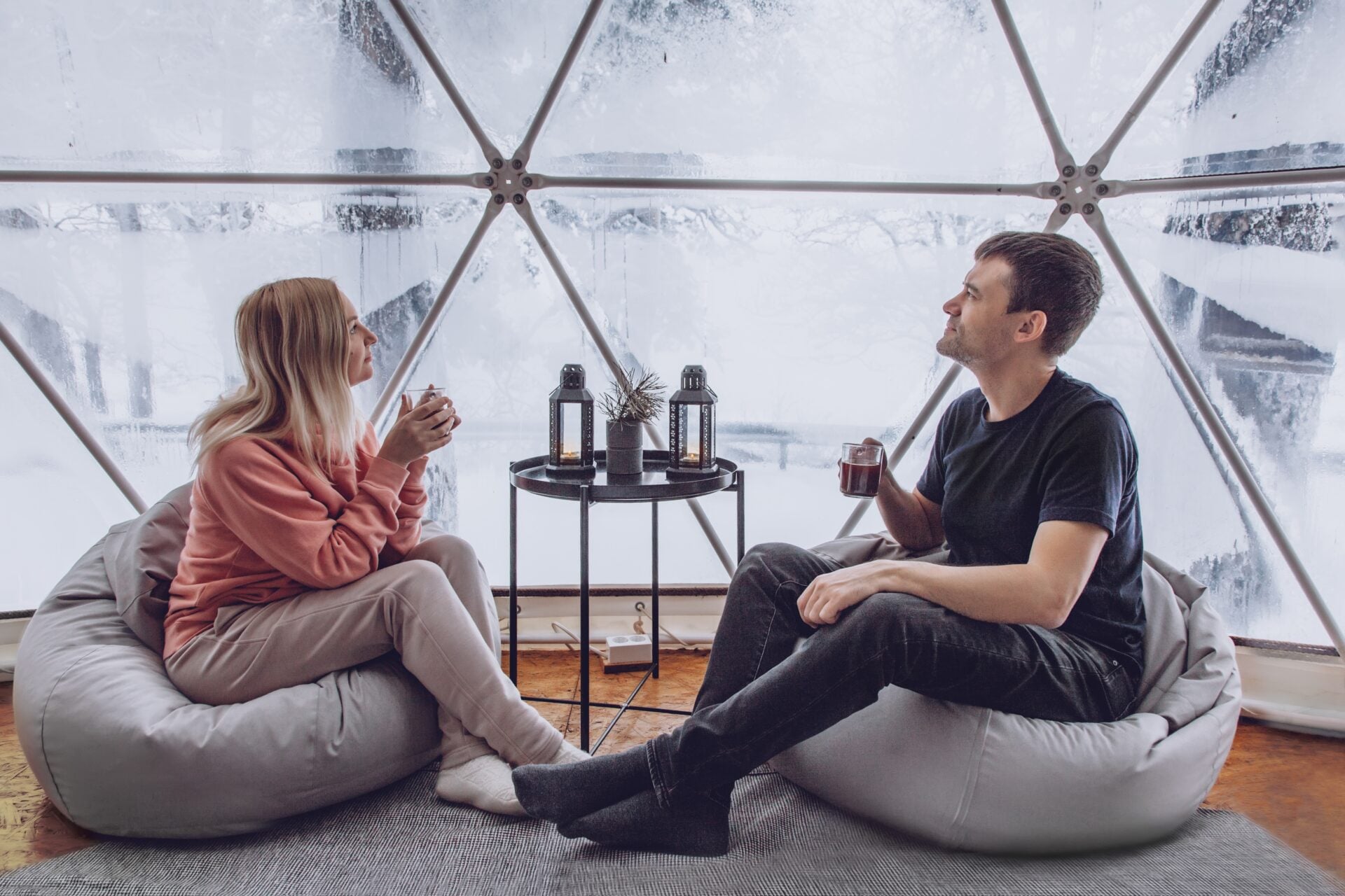 A couple - a man and a woman are sitting on bag chairs and looking out at the winter snow-covered nature and drinks coffee in the geodome of glamping.