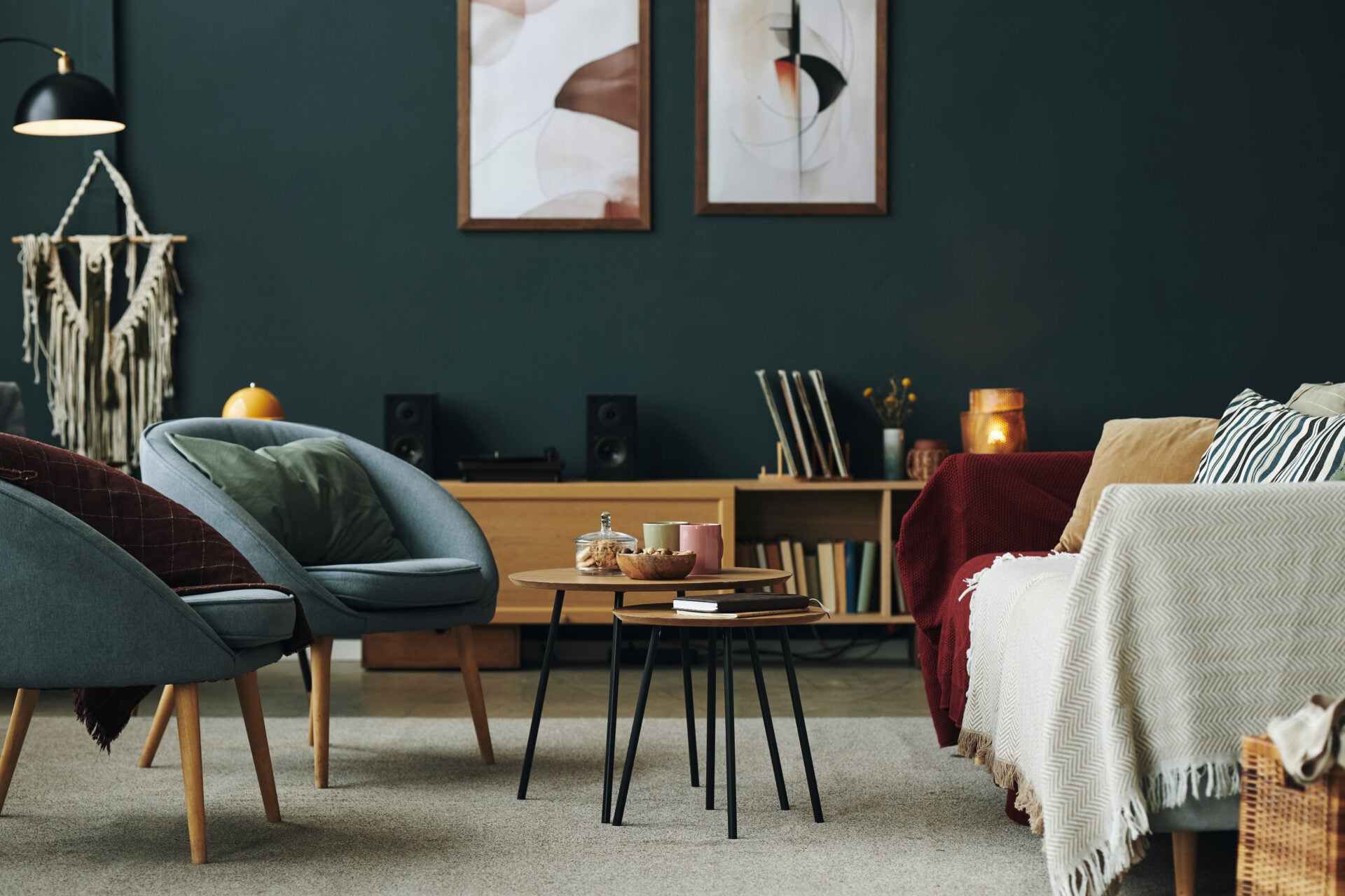 Modern living room featuring two armchairs and two sofas arranged around round coffee tables with books, candles, snacks, and abstract art on dark green wall in background