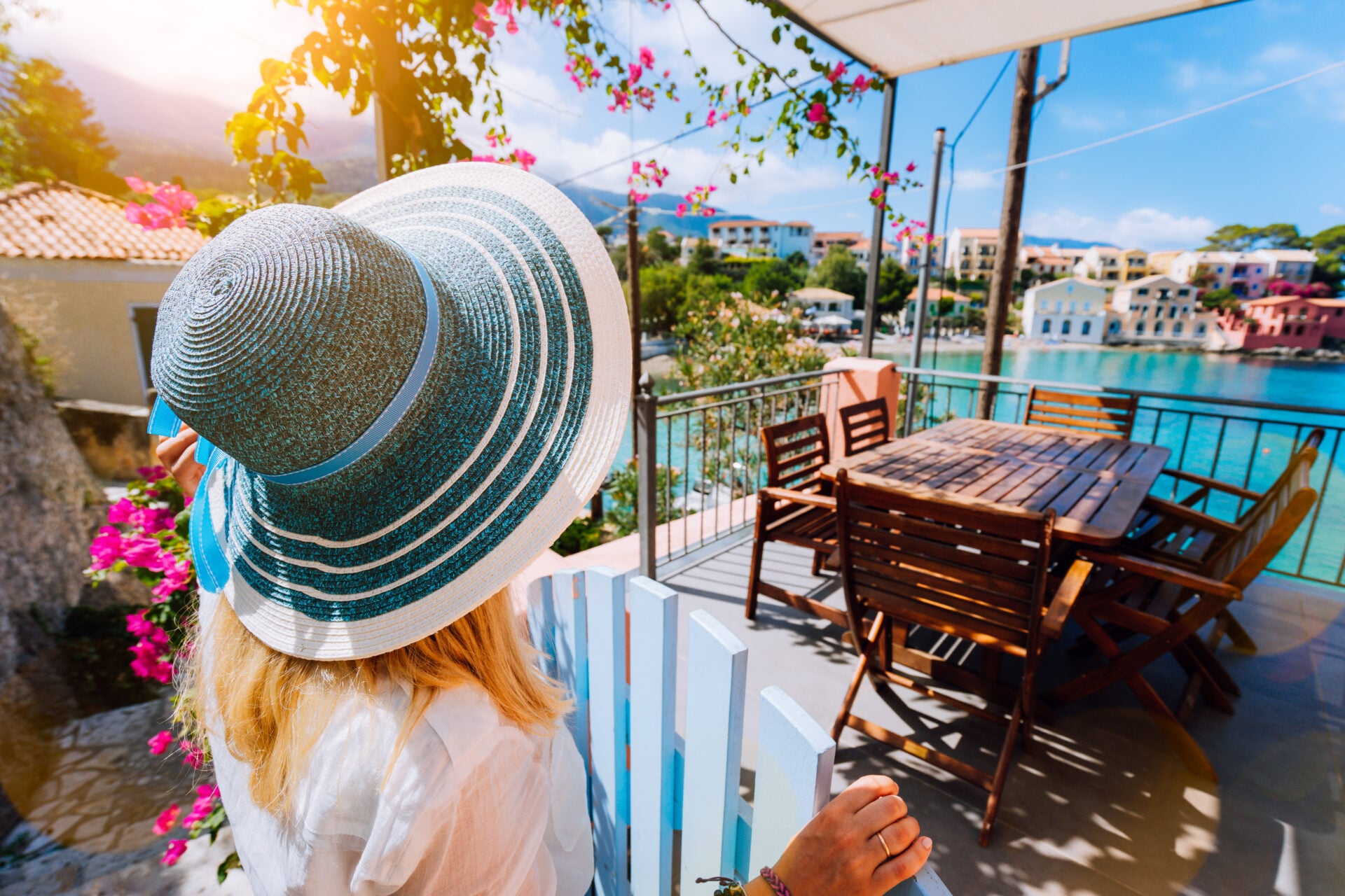 Female tourist in blue sunhat in Assos village in front of cozy veranda door admiring turquoise colored bay of Mediterranean sea and beautiful colorful houses of in Kefalonia, Greece.