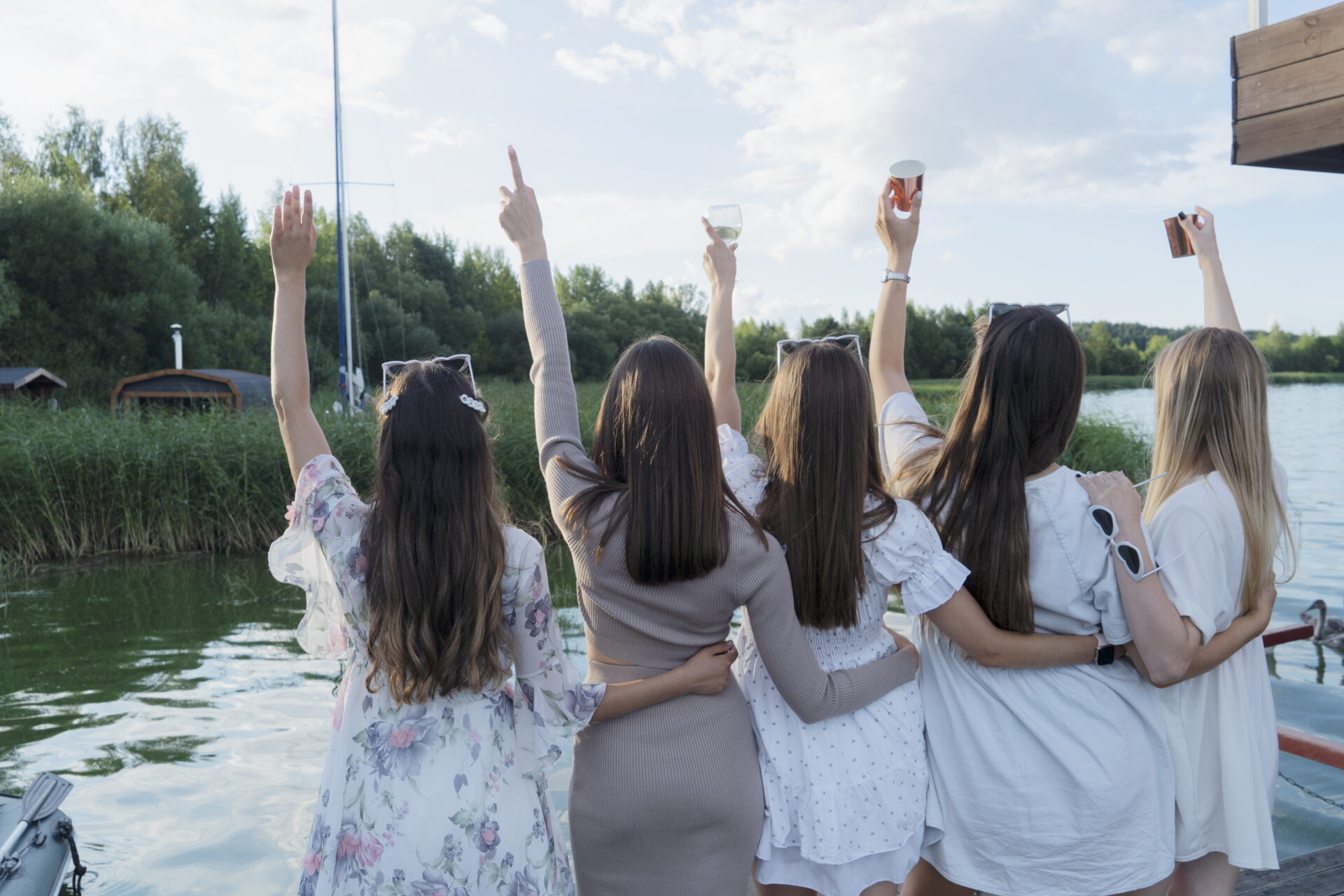 Five close friends gather at a lakeside, joyfully raising their drinks in cheers of friendship on a beautiful evening