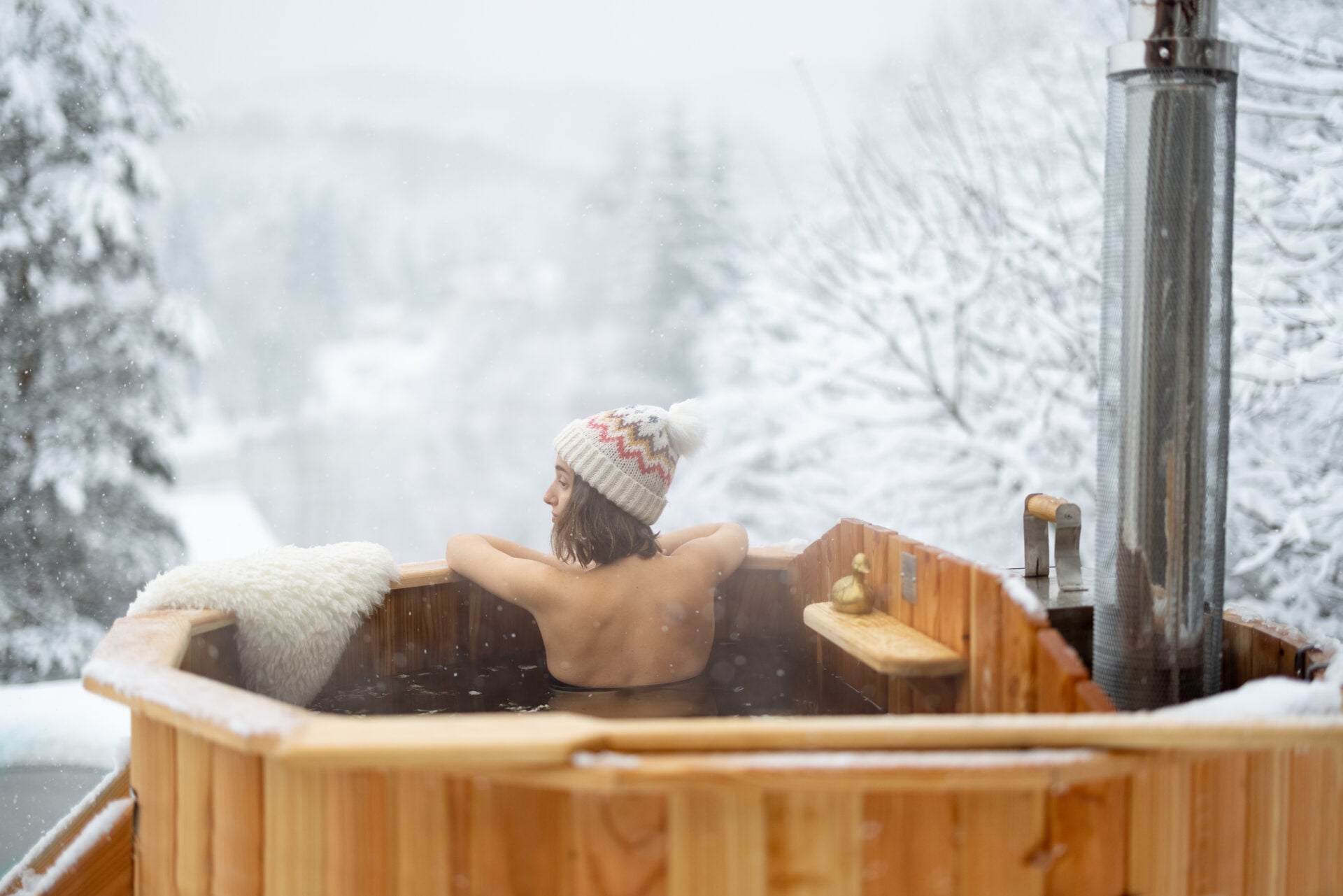 Woman relaxing in hot bath outdoors, sitting back and enjoying beautiful view on snowy mountains. Winter holidays in the mountains, hot water treatments concept. Caucasian woman wearing winter hat
