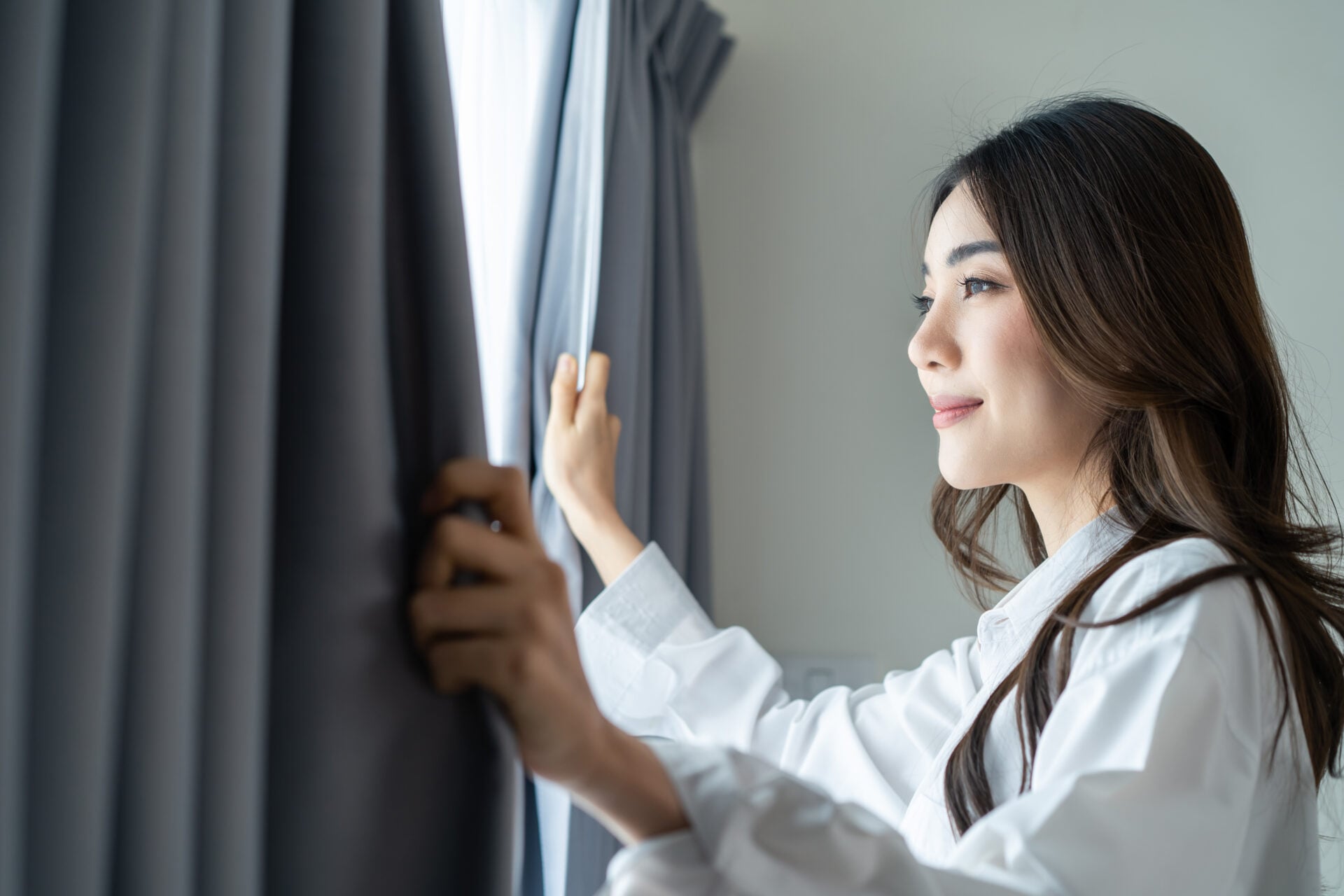 Asian beautiful woman approaches a window and open curtains in morning. Young attractive girl in white pajamas feel fresh and happy to look outside and sunrise after wake up early in bedroom at home.