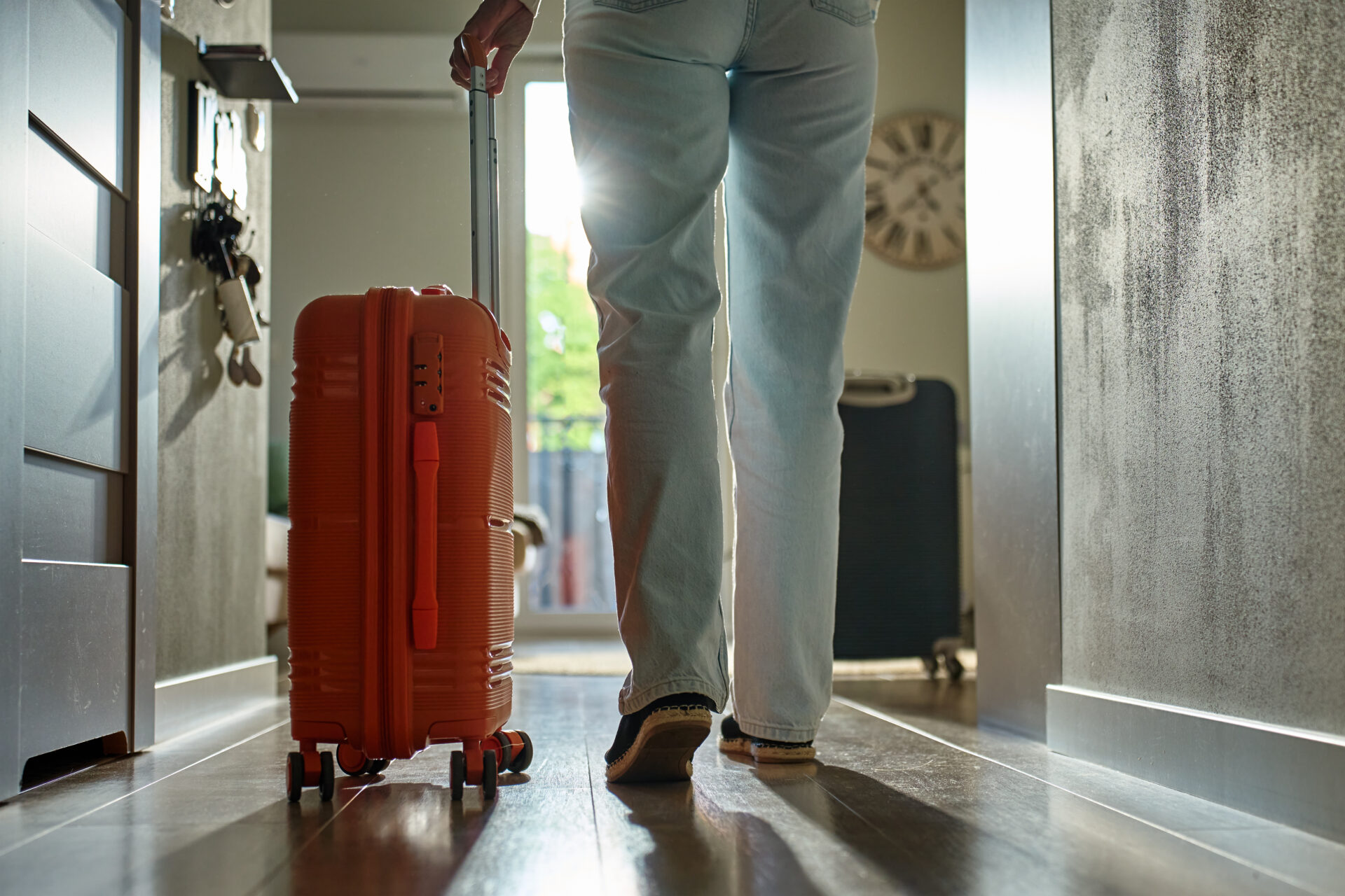 Low-angle view of traveler walking towards door with rolling luggage. Person walks across hallway with orange suitcase. Concept of departure and leaving home for vacation